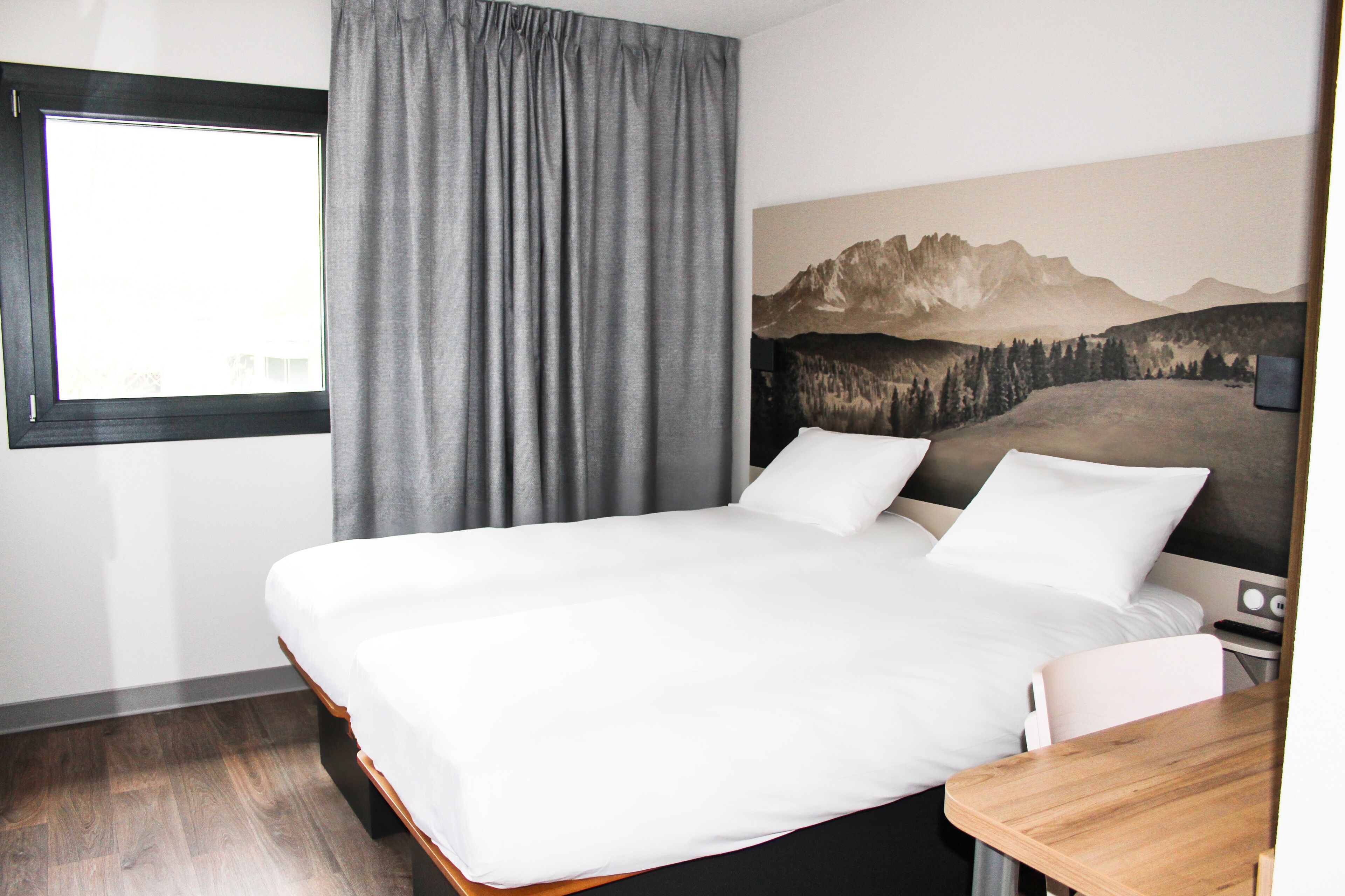 Quarto de hotel com uma cama grande, uma escrivaninha de madeira, uma janela com cortinas e uma pintura da paisagem de montanhas na parede.