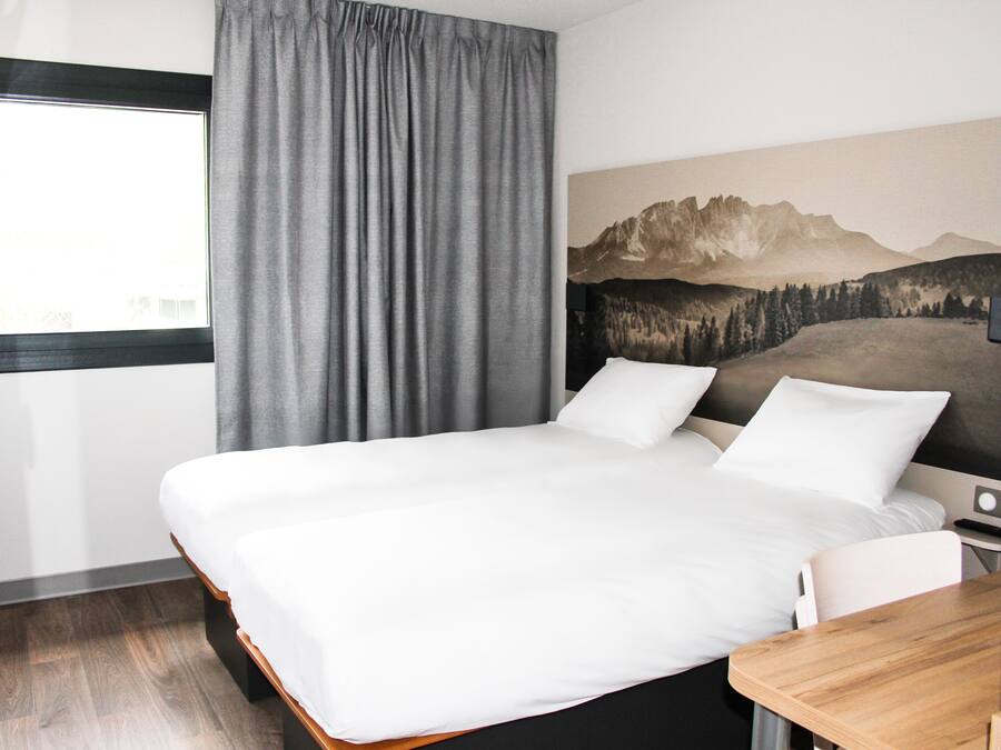 Quarto de hotel com uma cama grande, uma escrivaninha de madeira, uma janela com cortinas e uma pintura da paisagem de montanhas na parede.