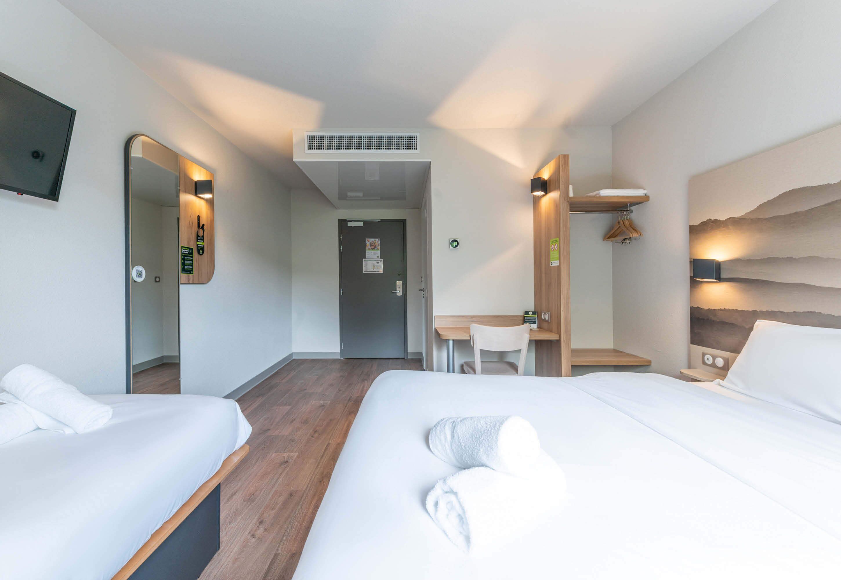 A hotel room with two beds, a desk, a chair, a mirror, and a wall-mounted TV.