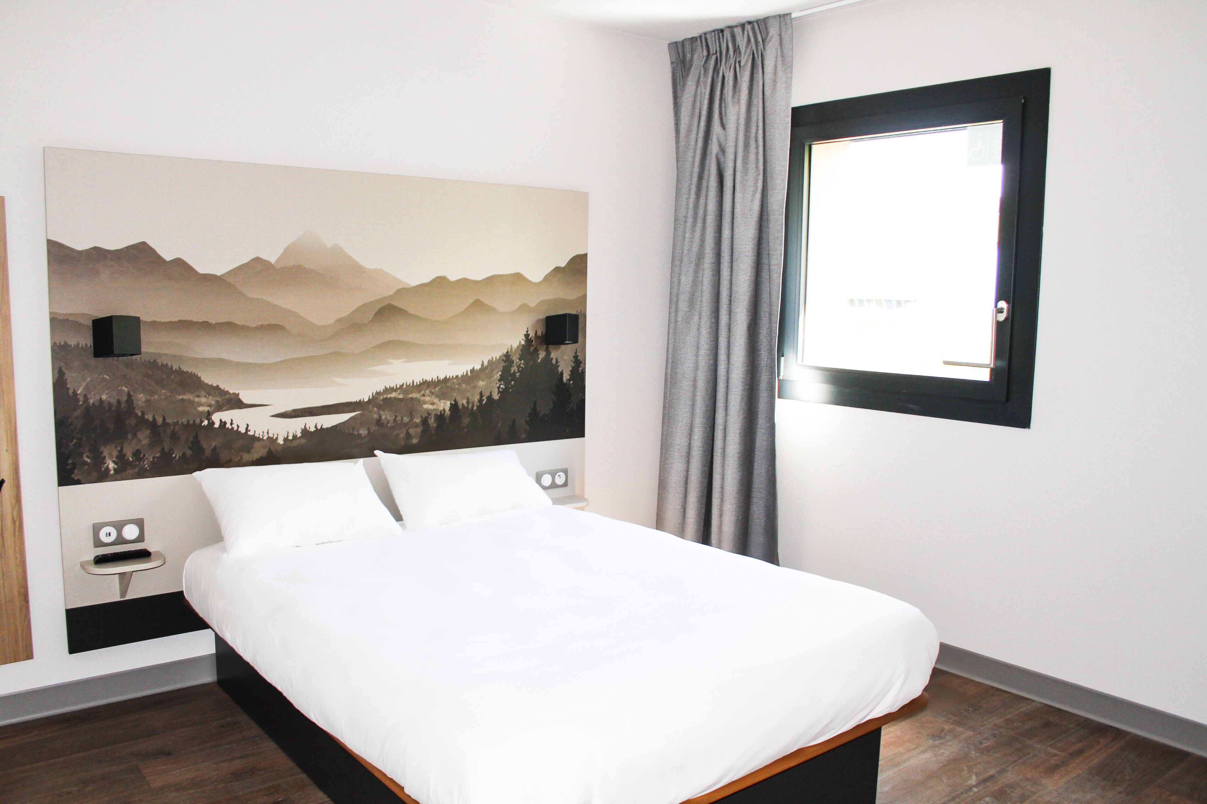A bedroom with a bed, a nightstand, a wall-mounted TV, a window with curtains, and a painting of a mountain landscape.