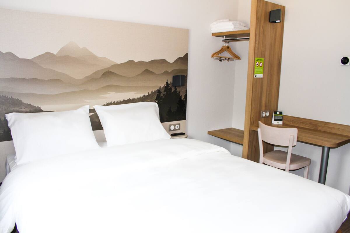Habitación de hotel con cama, escritorio, silla, televisor de pared y cuadro de montañas.