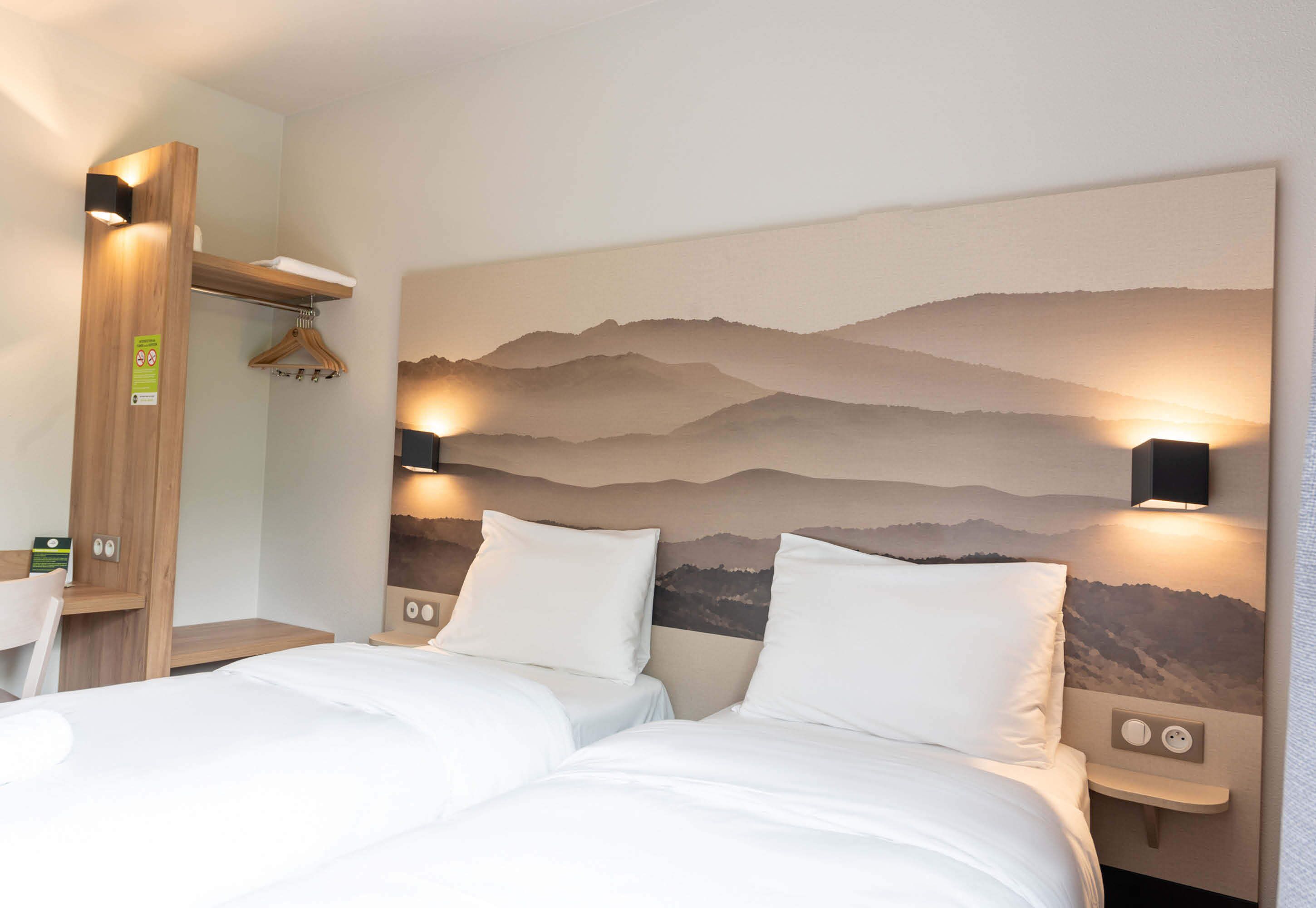 Een hotelkamer met twee bedden, een houten hoofdbord, een aan de muur bevestigde televisie en een berglandschap op de muur.