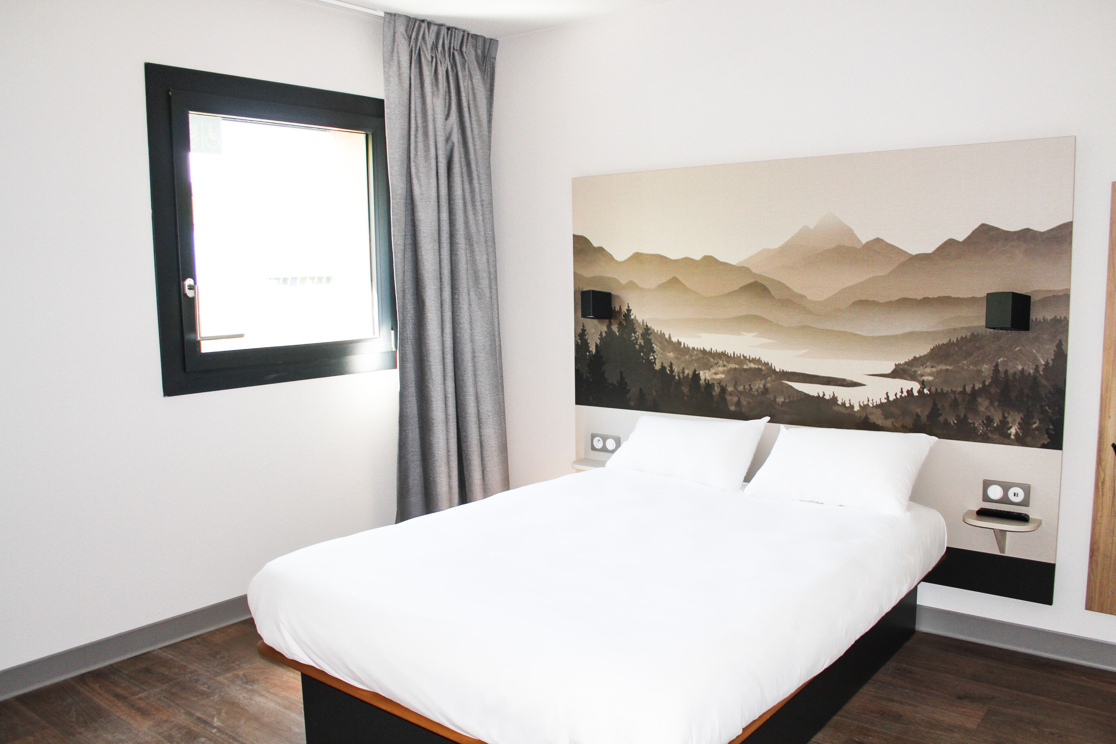 Un dormitorio con cama, ventana con cortinas, una obra de arte en la pared que representa un paisaje montañoso y una mesita de noche con lámpara.