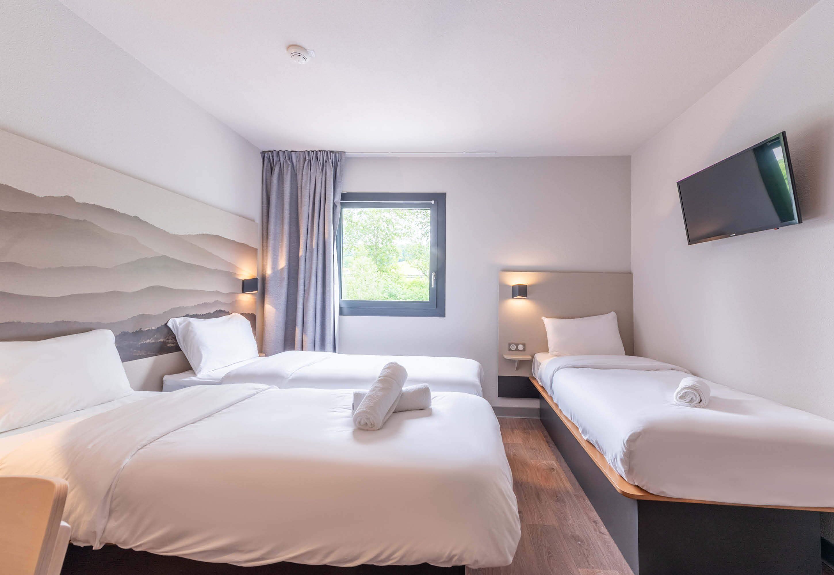A hotel room with two beds, a large window, a flat-screen TV, and a wall-mounted light fixture.