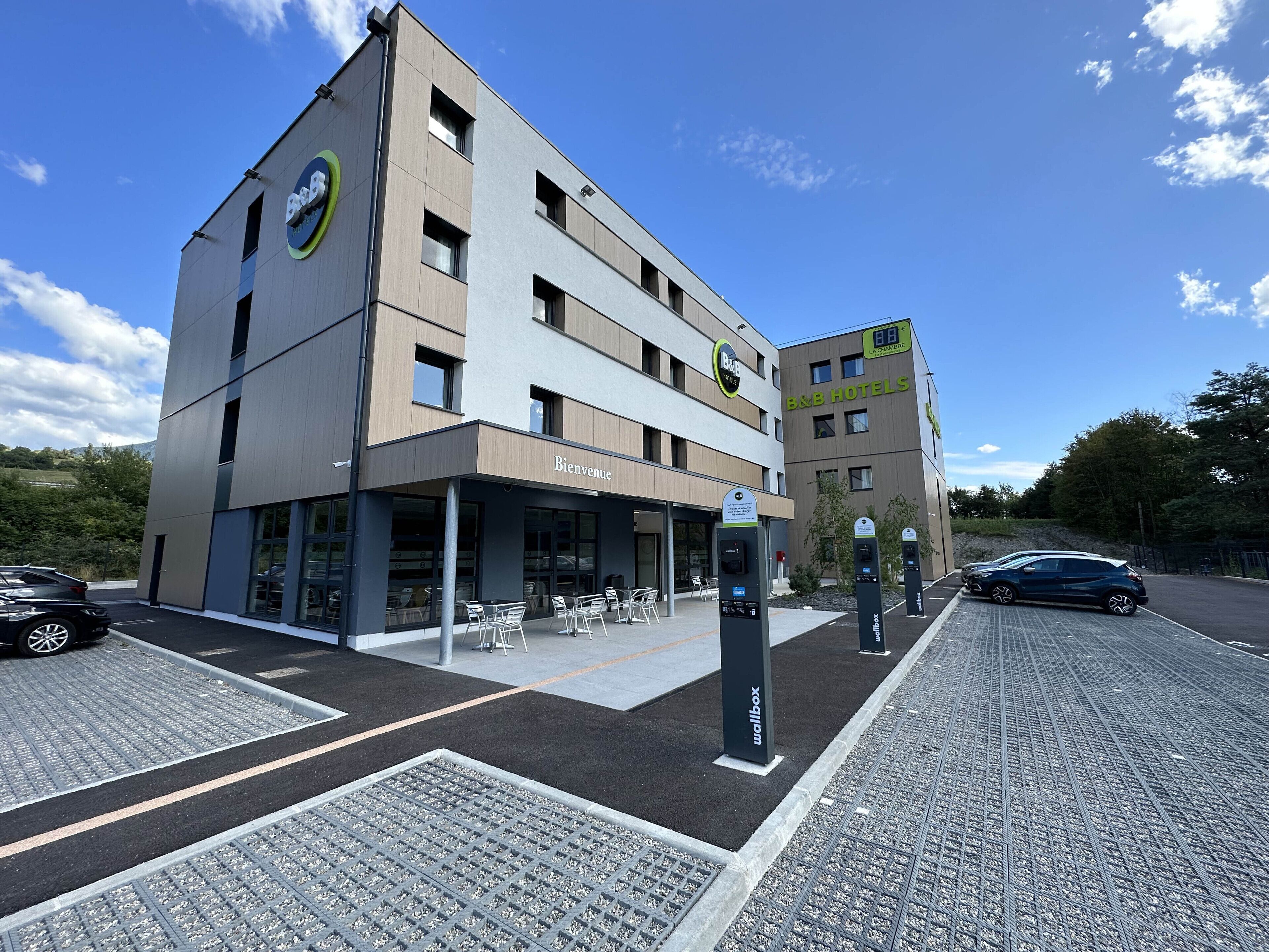 Un hotel moderno con un cartello 'Bienvenue', area con posti a sedere all'aperto e parcheggio dotato di stazioni di ricarica per veicoli elettrici.