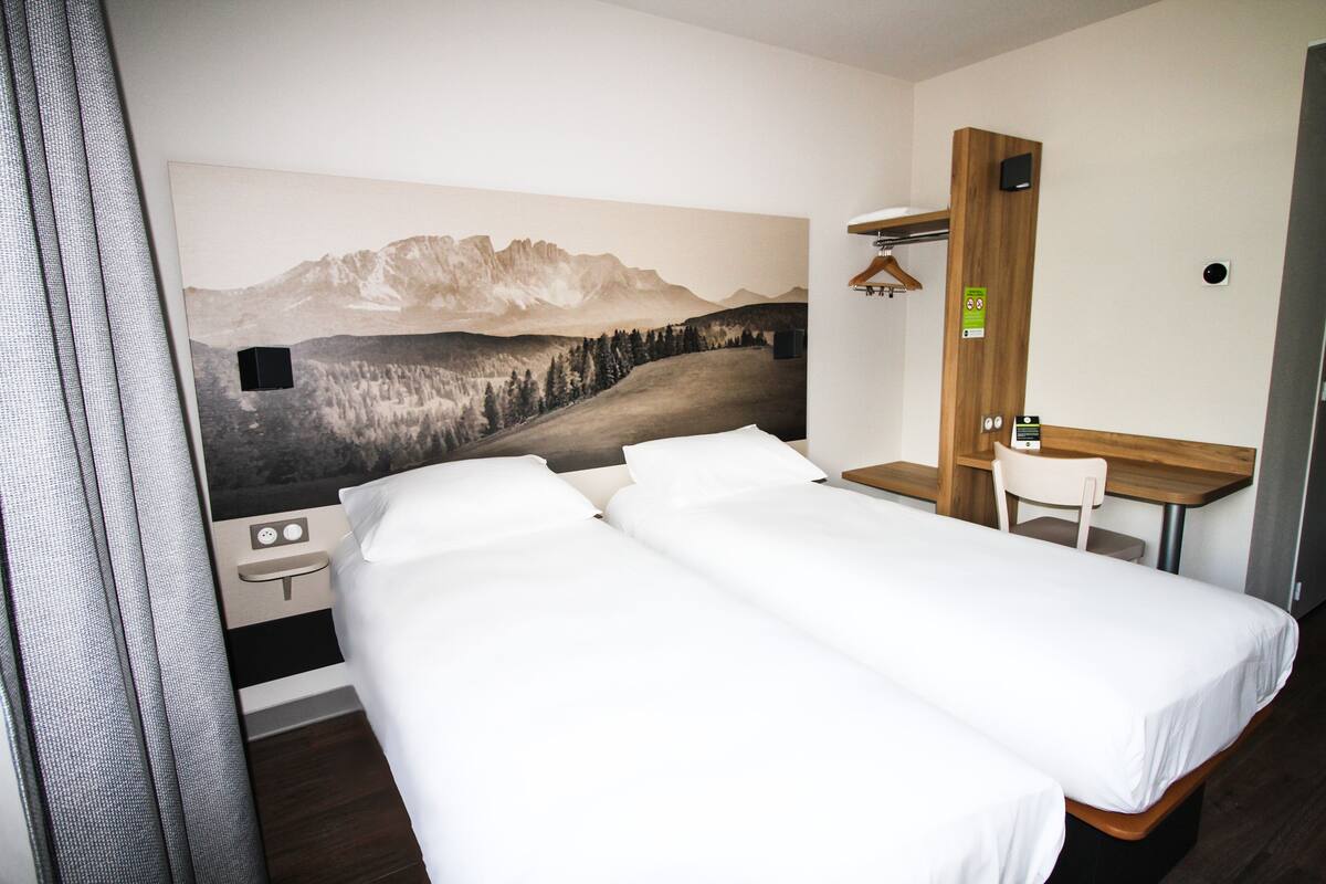 Habitación de hotel con dos camas, un escritorio, una silla y un mural con paisaje de montaña en la pared.