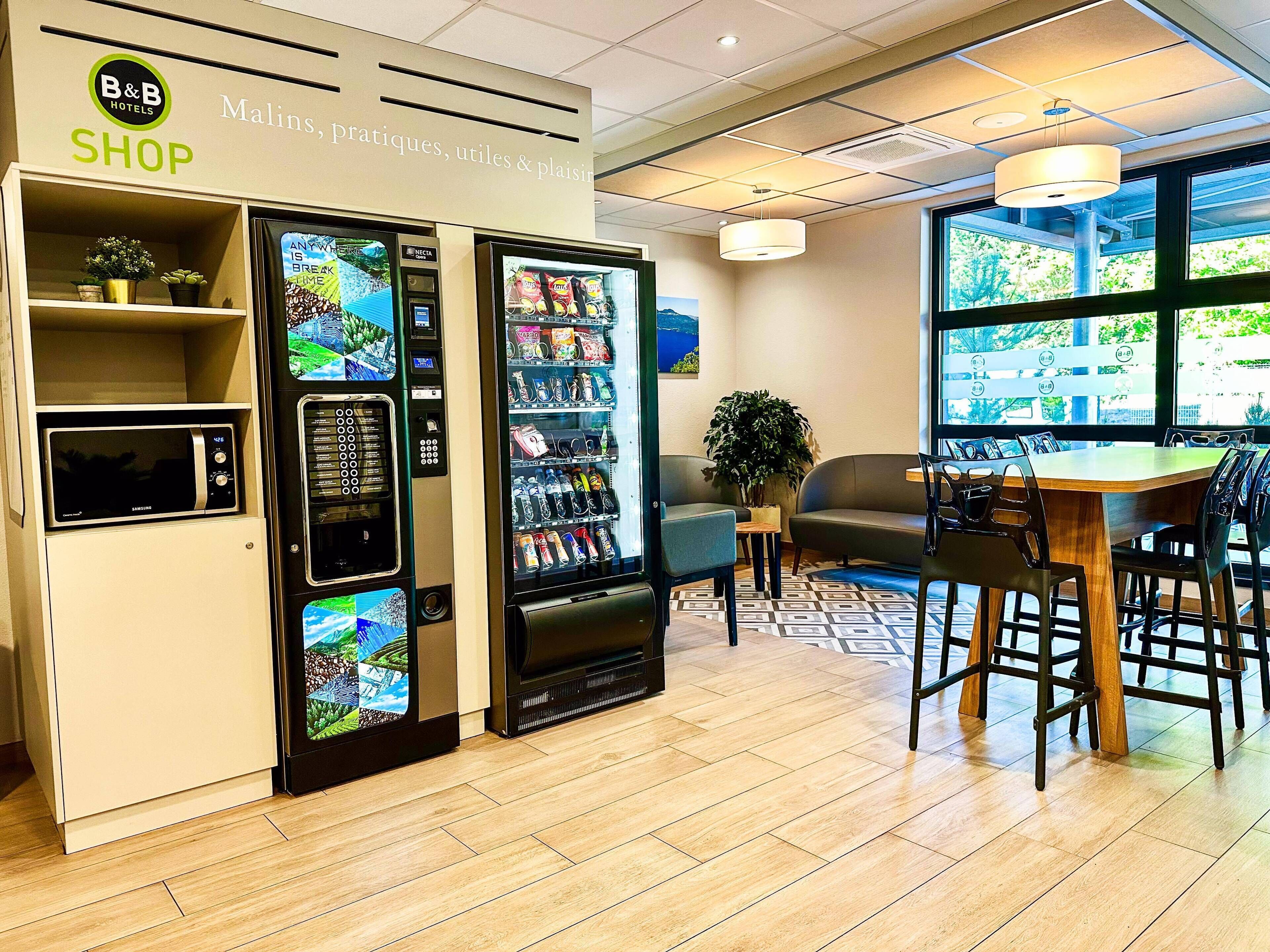 Un magasin d’hôtel de type « bed and breakfast » avec des distributeurs automatiques, un coin salon avec une table et une vue sur la verdure environnante.