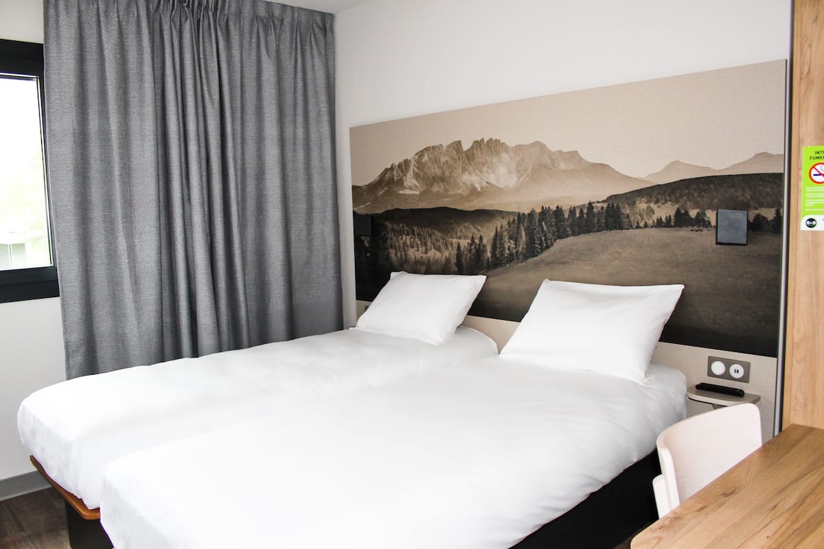 Habitación de hotel con cama, escritorio, silla, ventana con cortinas y televisión instalada en la pared.