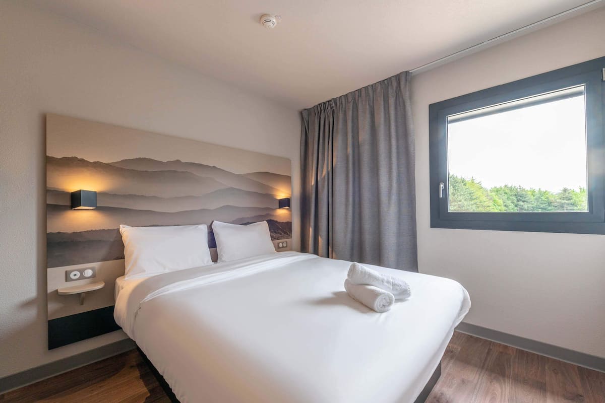 Una cama bien hecha con una toalla doblada, un cabecero con un cuadro iluminado en la pared, una mesita de noche y una ventana con cortinas.