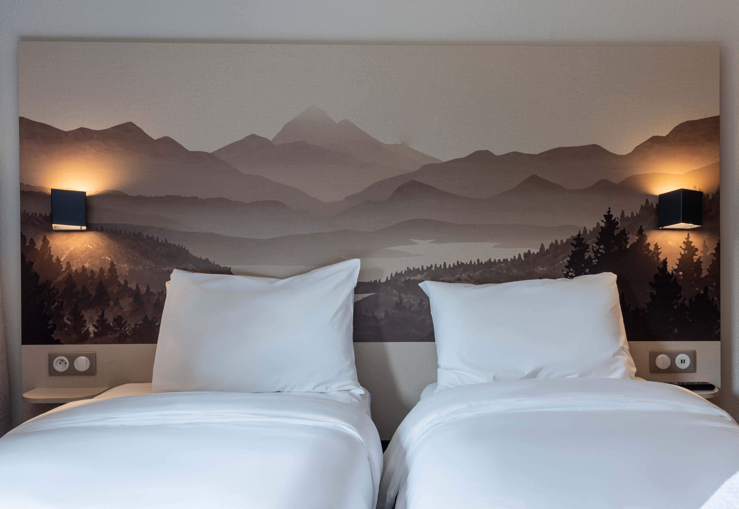 A hotel room with two beds, a mountain landscape mural on the wall, and two wall-mounted lamps.