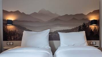 A hotel room with two beds, a mountain landscape mural on the wall, and two wall-mounted lamps.