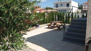 A patio area with a wooden table and benches, surrounded by olive trees and potted plants.
