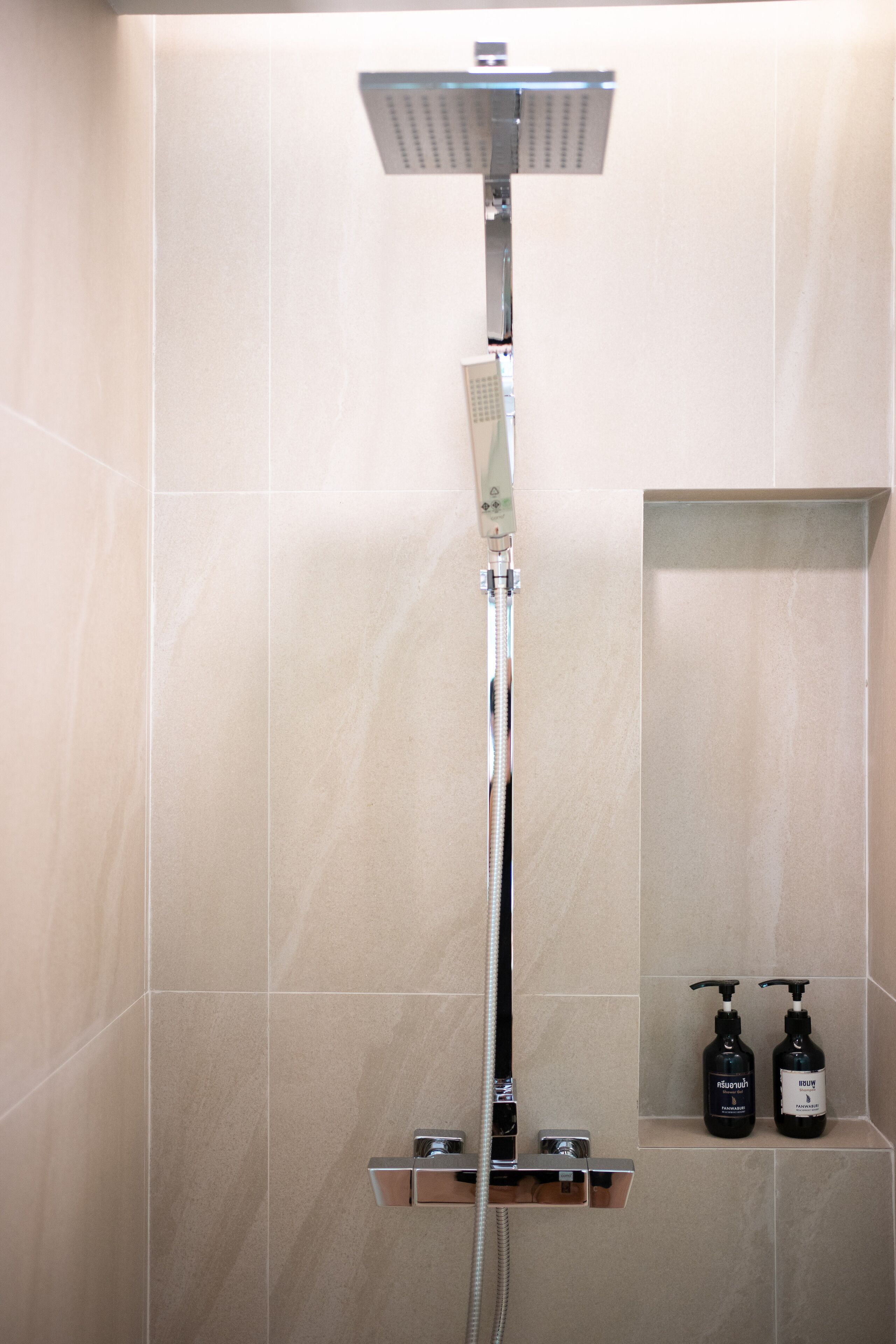 A shower with a handheld showerhead and a wall-mounted control panel. There are two bottles of shampoo and conditioner on a shelf below the shower.