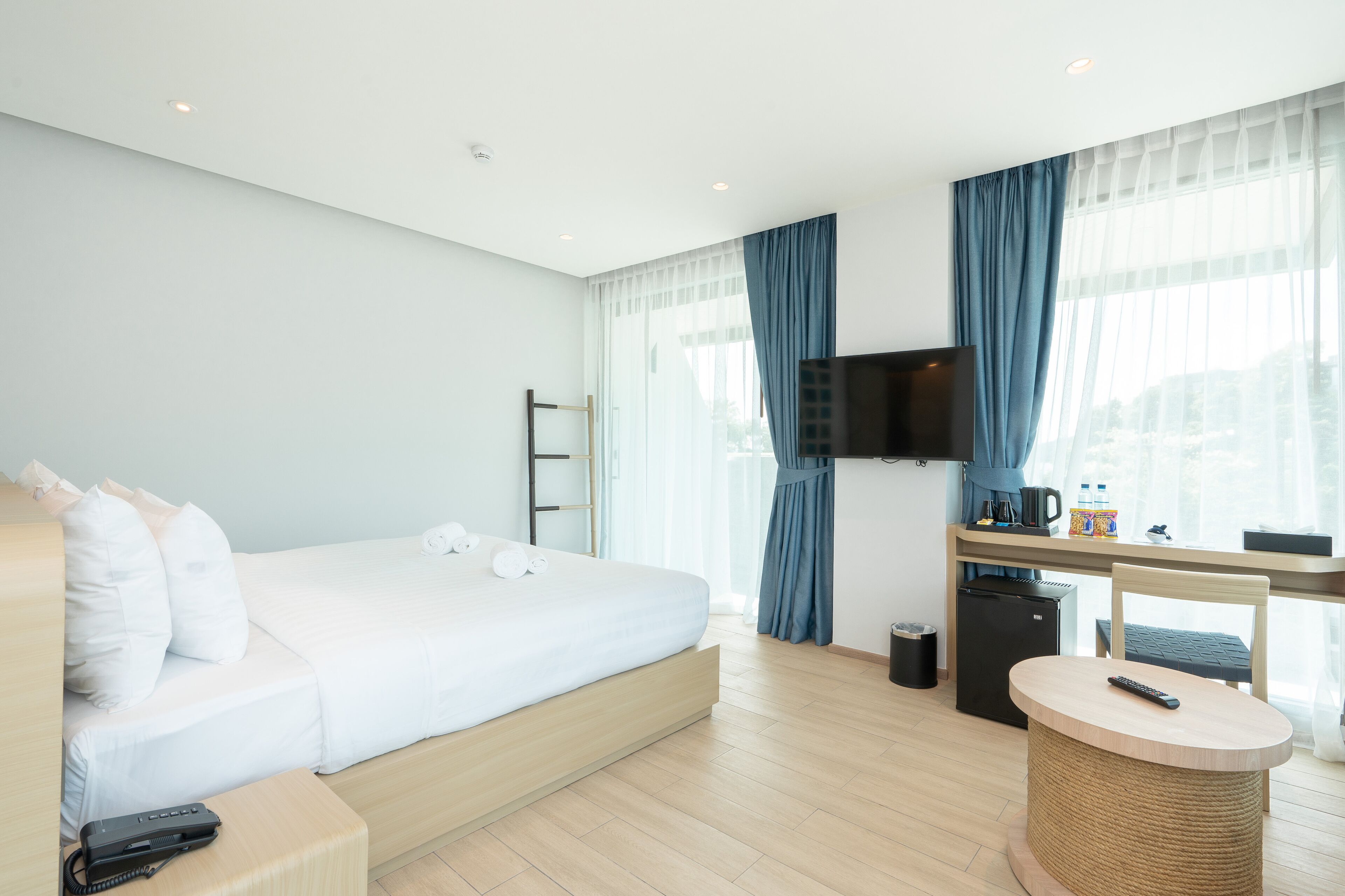 Una habitación de hotel moderna con una cama grande, un escritorio con televisor, un refrigerador pequeño y una mesa redonda con un control remoto.