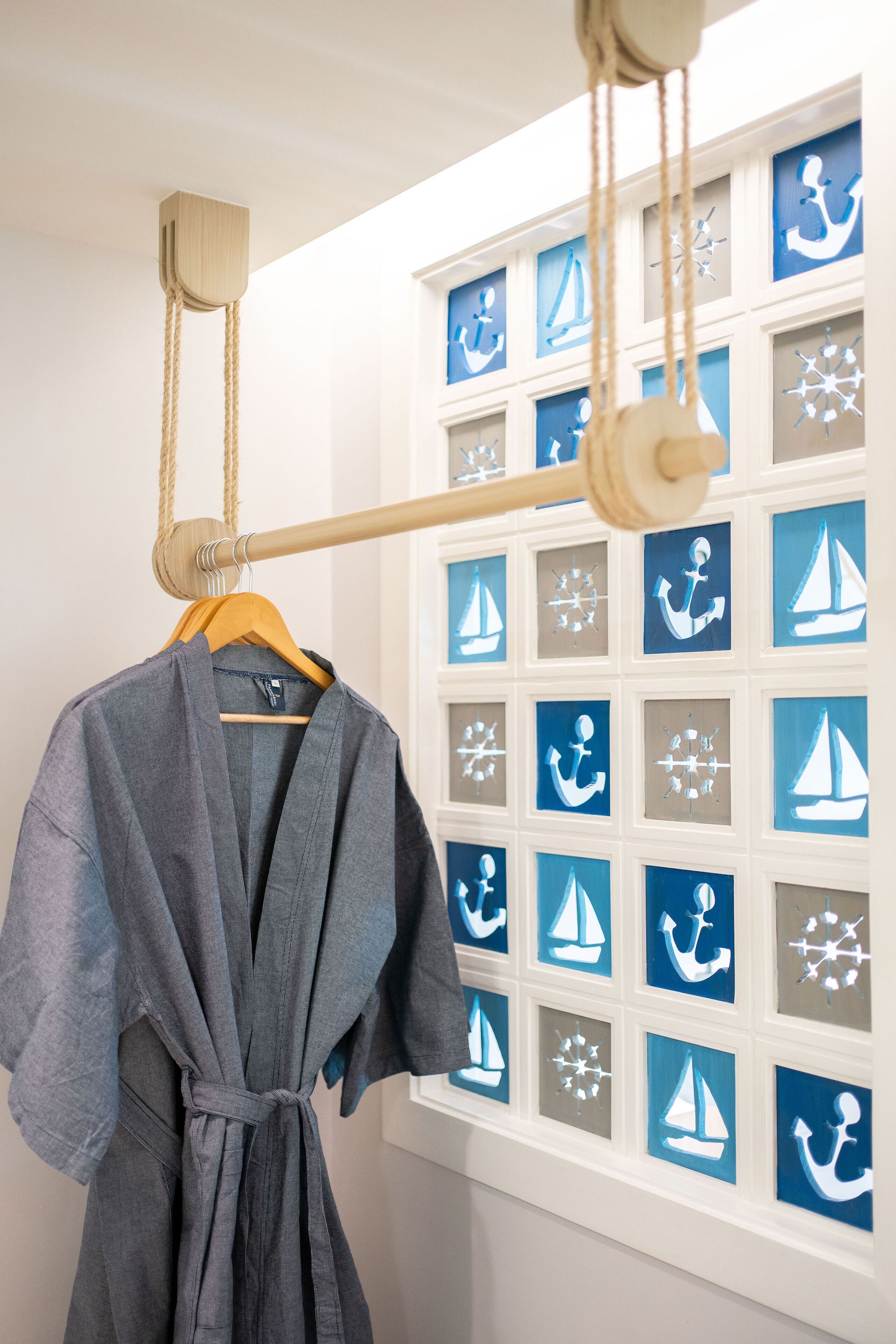 A bathroom with a wooden towel rack holding a grey robe, a window with nautical-themed artwork, and a white wall.