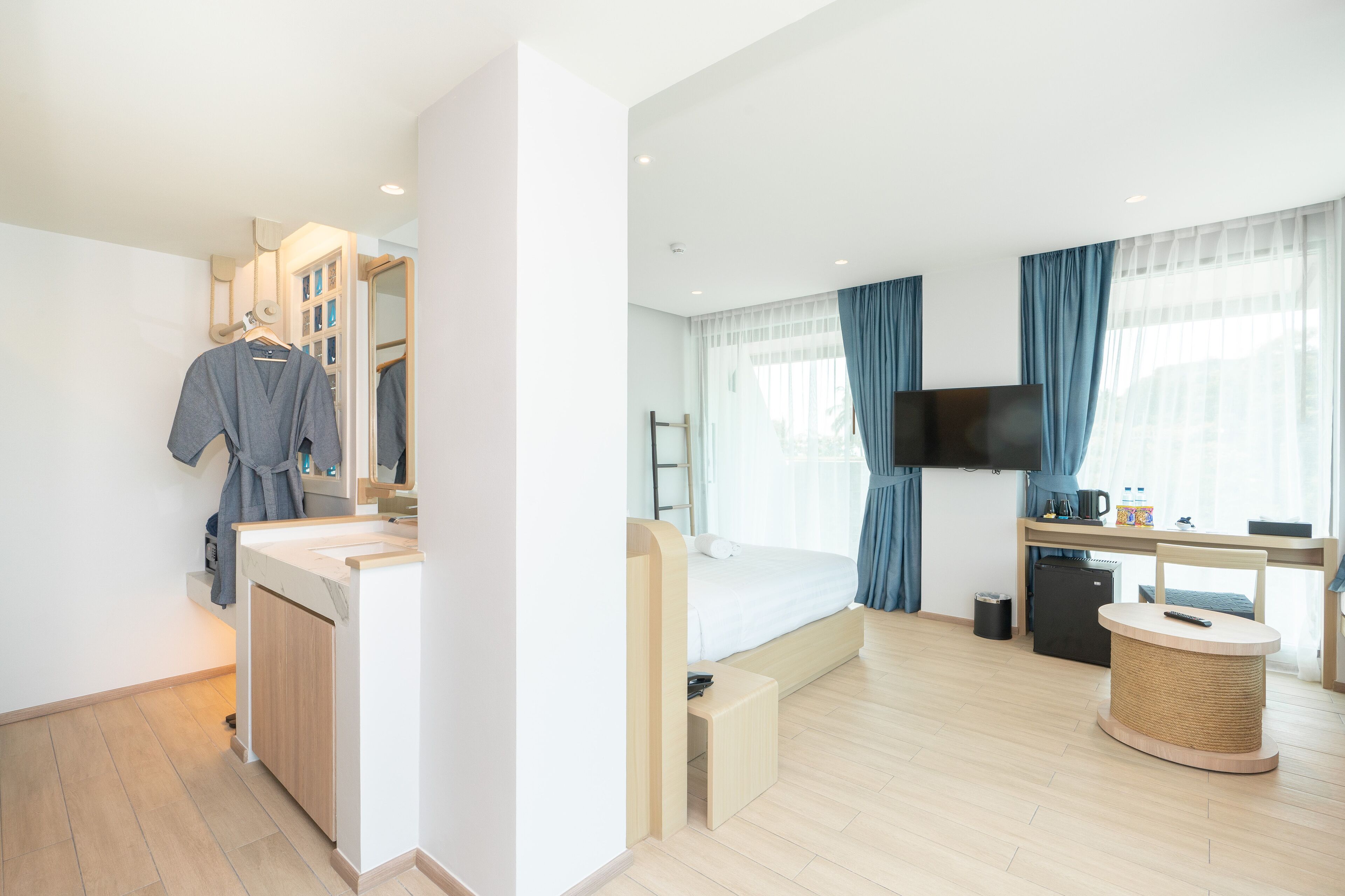 A modern hotel room with a bed, a TV, a desk, and a bathroom with a mirror and towel rack.