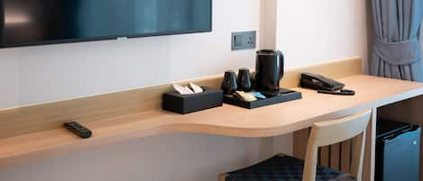 A modern hotel room with a flat-screen TV mounted on the wall, a wooden desk with a coffee maker, a phone, and a remote control. There is a chair with a woven seat and a black trash bin on the floor.