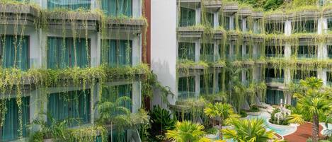 Un hotel con varios balcones cubiertos de vegetación, una piscina central rodeada de palmeras y un paisaje exuberante al fondo.
