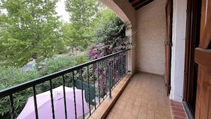 Un balcón techado con suelo de baldosas y barandilla de metal, con vista a un jardín con diversas plantas y un arbusto de flores moradas.