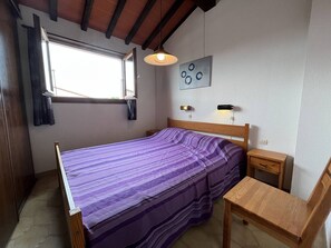 A bedroom with a bed, a wooden nightstand, a small table, a hanging light fixture, and a window with curtains.
