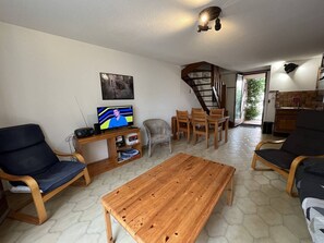 A living room with a wooden coffee table, two armchairs, a TV, and a bookshelf. A staircase leads to a kitchen area.