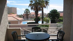 A balcony with a round table and four chairs, overlooking a swimming pool and residential buildings. - Cozy 1-Bedroom Apartment with Pool, Near Beach & Port, Sleeps 5, Pet-Friendly! (Saint-Cyprien)