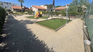 A playground with a slide and swings, surrounded by a fence and greenery. - Cozy 1-Bedroom Apartment with Pool, Near Beach & Port, Sleeps 5, Pet-Friendly! (Saint-Cyprien)