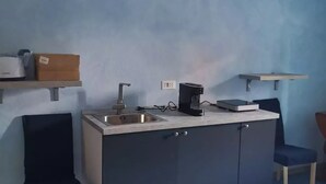 A compact kitchenette with a sink, a coffee machine, and a small countertop. There are two chairs and a shelf above the counter. - Guesthouse La nuvola (Sagliano Micca)