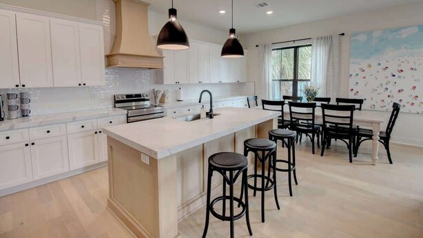 A modern kitchen with white cabinetry, a central island with stools, pendant lighting, and a dining area with black chairs. - Galveston Modern - 4BR with Panoramic Views (Galveston)