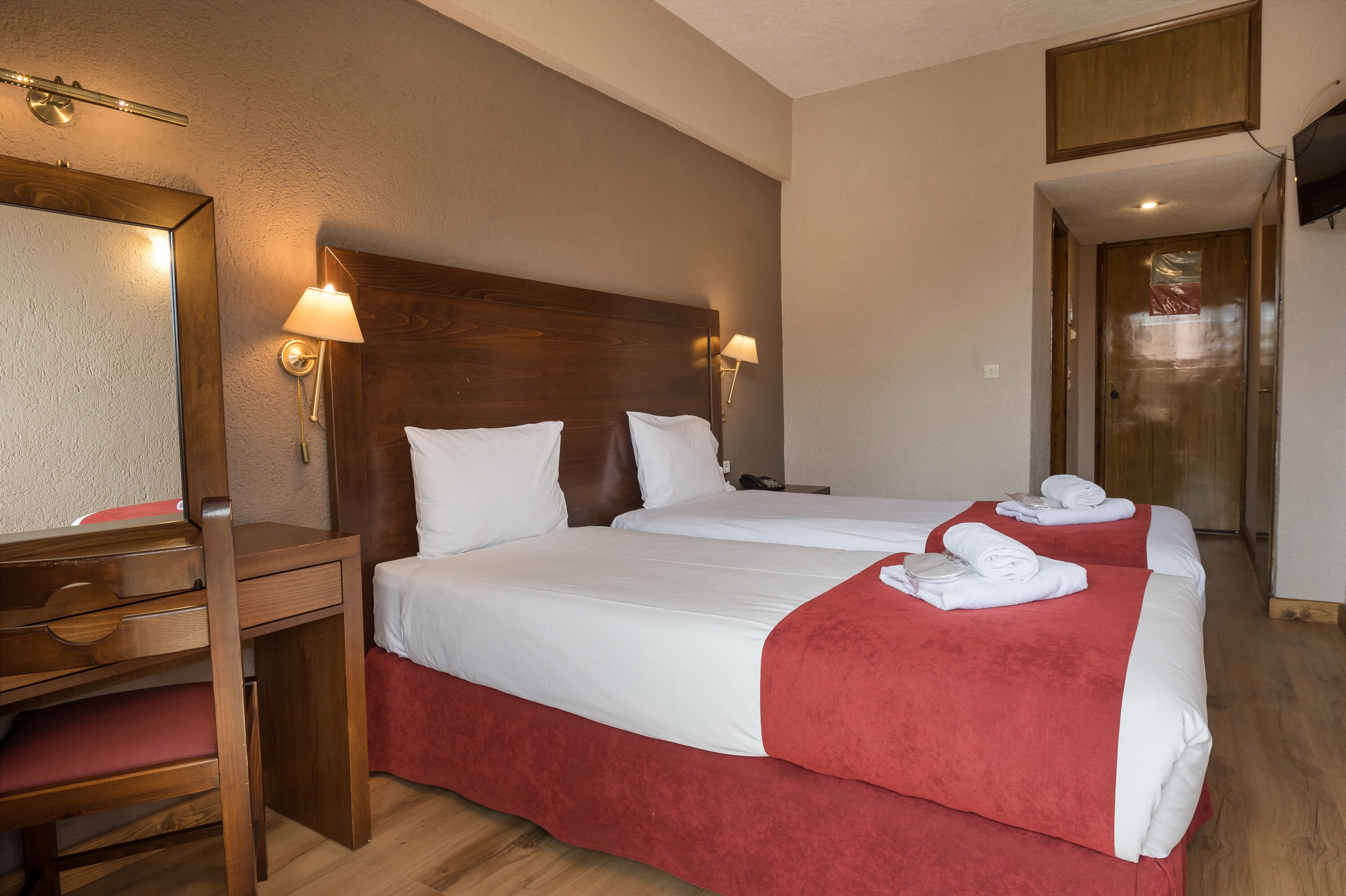 A hotel room with two beds, a wooden desk, a mirror, and two wall-mounted lamps.