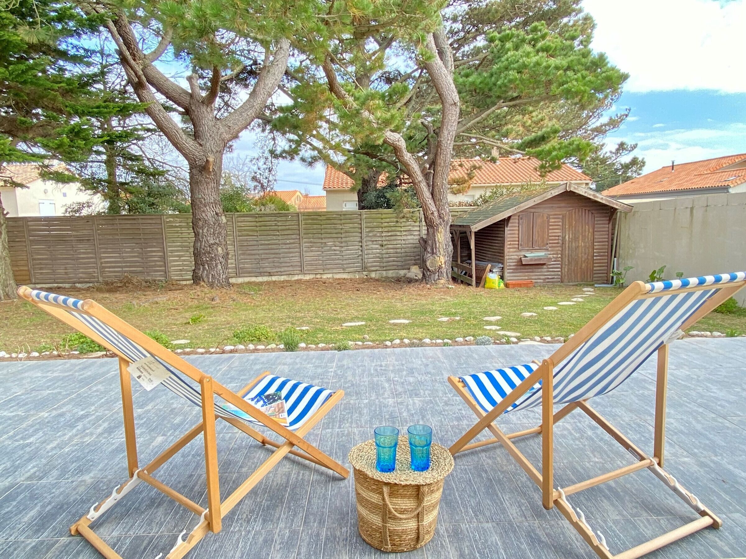 Deux chaises de plage à rayures, séparées par un panier en osier, sont placées sur une terrasse. On aperçoit une clôture en bois et des arbres en arrière-plan.