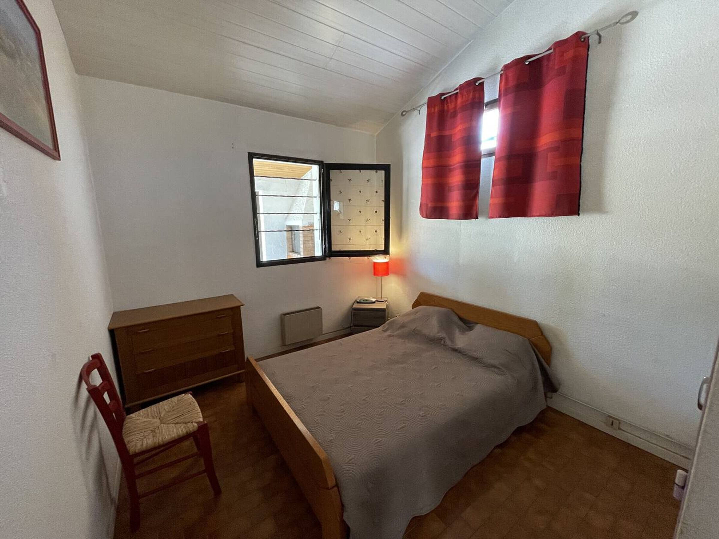 Ein kleines Schlafzimmer mit einem Bett, einer Holzkommode, einem Stuhl, einem Nachttisch mit Lampe, einem Fenster mit Vorhängen und einem Heizkörper.
