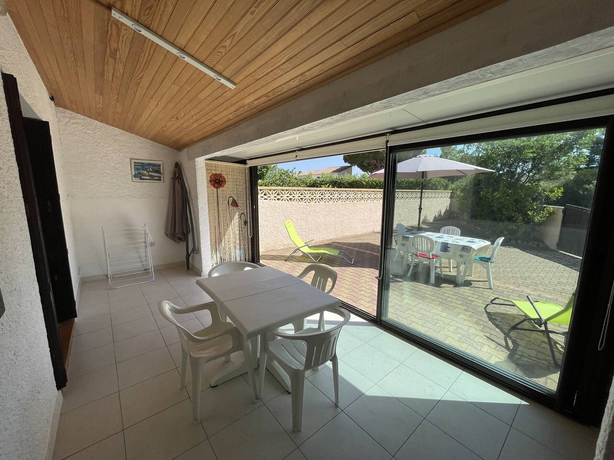 Een kamer met een houten plafond, witte wanden, een betegelde vloer, een tafel met stoelen, en een glazen schuifdeur die leidt naar een buiten terras met een tafel en stoelen.