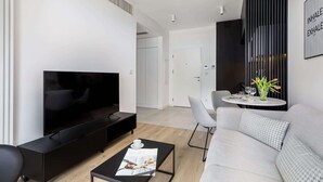 Ein modernes Wohnzimmer mit Flachbildfernseher, einer Couch, einem Couchtisch mit einer Zeitschrift und einem Essbereich mit kleinem Tisch und Stühlen.