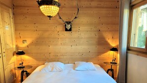 A wooden room with a bed, two lamps, a chandelier, and a deer skull on the wall.