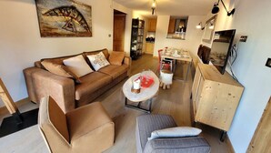 A cozy living room with a brown sofa, a small round table, and a grey armchair. There is a wooden cabinet and a painting on the wall.