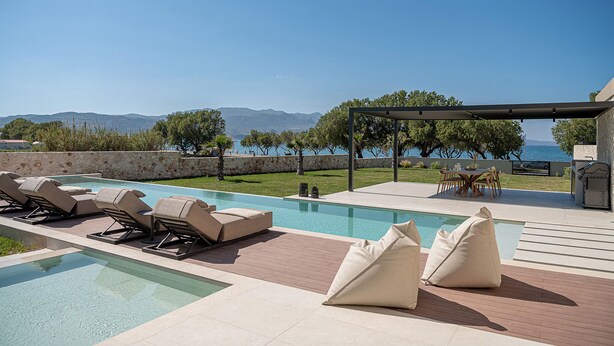 Zona de piscina con sillas de descanso y una mesa, con vistas a un cuerpo de agua y montañas.