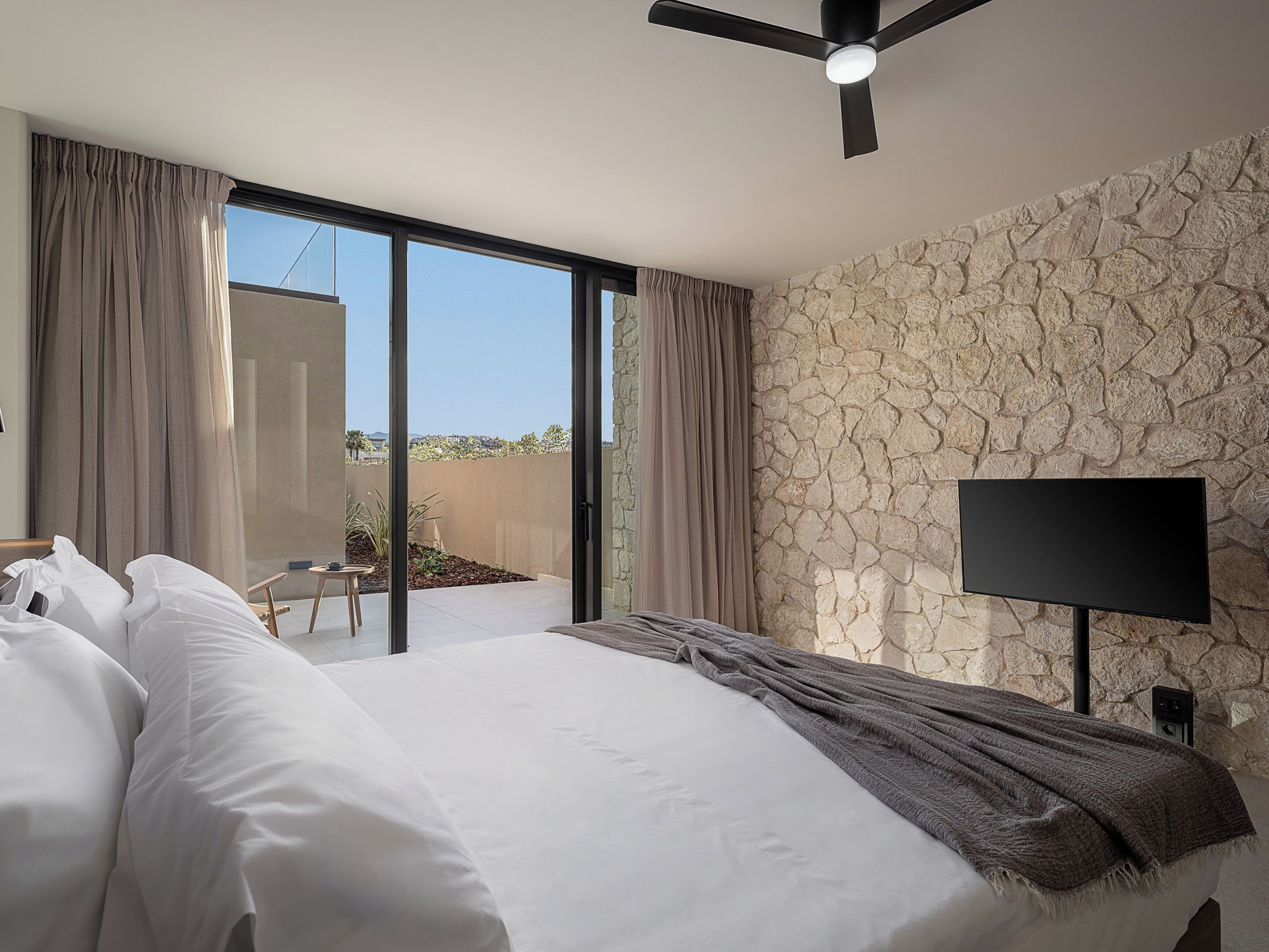 A modern hotel room with a large bed, a flat-screen TV, a ceiling fan, and a view of the outdoors.