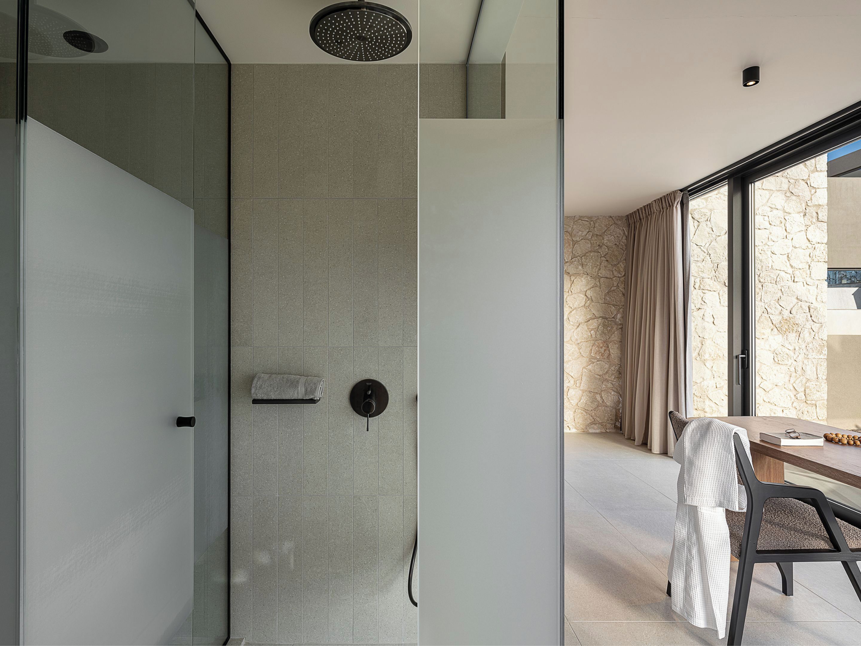 A modern bathroom with a glass shower enclosure, a towel rack, and a stone wall visible through a window.