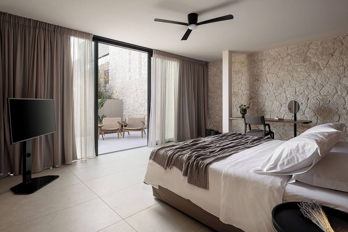 A modern hotel room with a bed, a flat-screen TV, a ceiling fan, and a stone-textured wall.