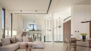 Amplia sala de estar con un sofá seccional grande, una mesa de centro redonda y una mesa de comedor con sillas. Una escalera conduce a un nivel superior, y la habitación cuenta con ventanas grandes que permiten la entrada de luz natural.