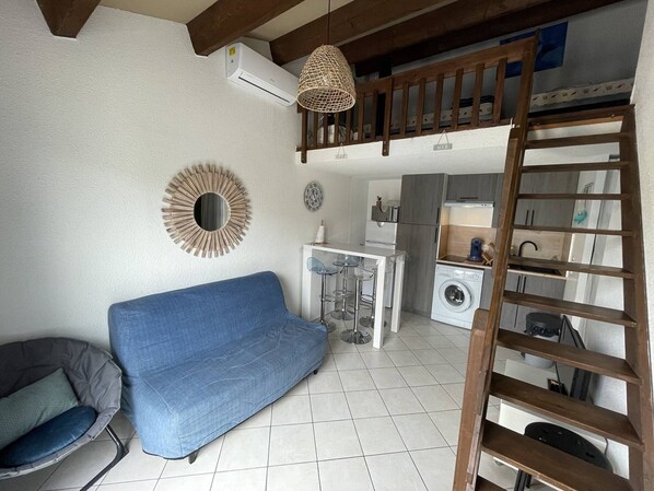 Un espacio habitable compacto con sofá, kitchenette pequeña, lavadora y un altillo con escalera.
