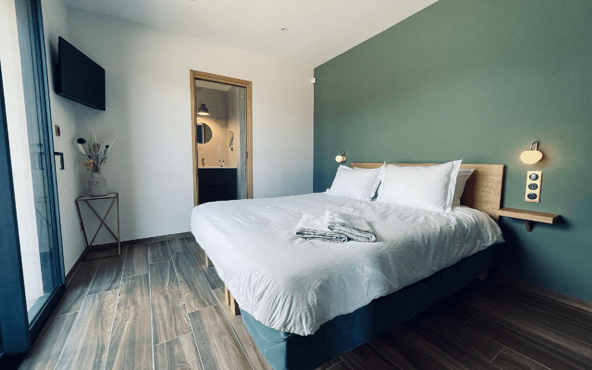 A modern bedroom with a large bed, a bedside table, a mirror, and a wall-mounted light fixture.