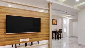 A modern living room with a flat-screen TV mounted on a wooden panel, a dining area with a table and chairs, and a hallway leading to another room. - Click Collection Jaipur (Jaipur)