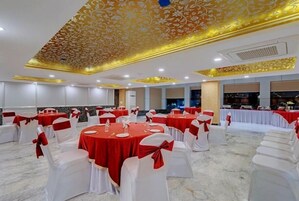 Salão de banquetes com mesas redondas cobertas por toalhas vermelhas e cadeiras brancas com fitas vermelhas. O teto apresenta um padrão floral com detalhes dourados.