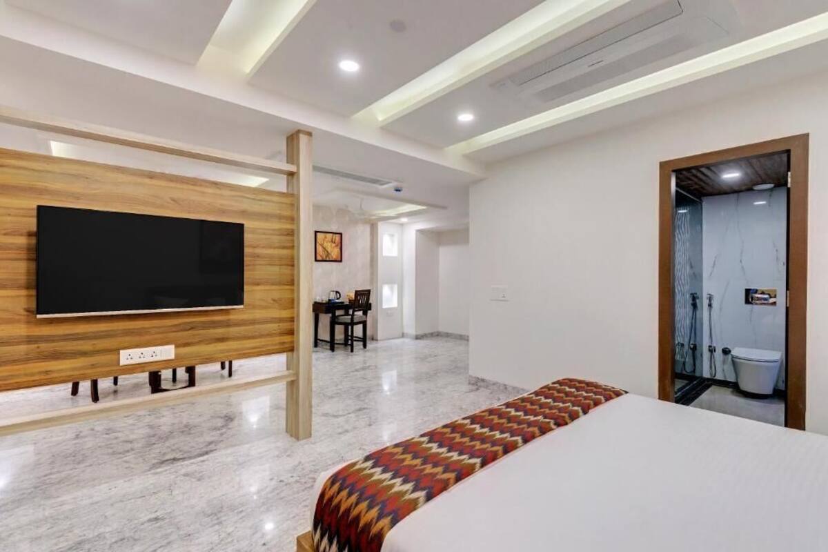 Ein modernes Hotelzimmer mit einem großen Flachbildfernseher, der an einer Holzvertäfelung befestigt ist, einem Bett mit einem gemusterten Überwurf, einem kleinen Tisch mit Stühlen und einem durch eine offene Tür sichtbaren Badezimmer.