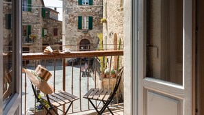 Ein Balkon mit Korbstühlen und einem Holztisch, mit Blick auf einen Innenhof mit Steingebäuden und grünen Fensterläden.