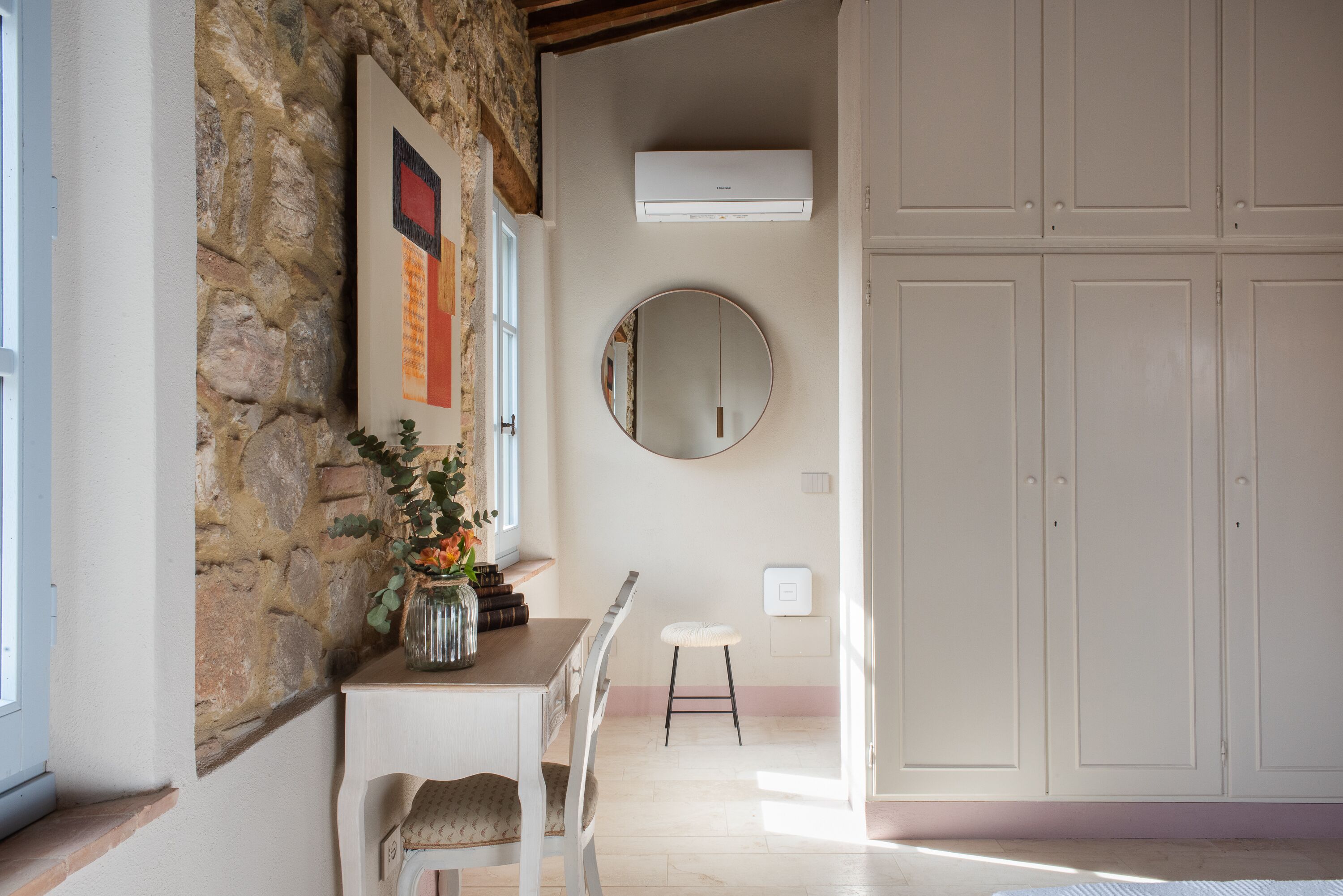 Una stanza con parete in pietra, uno specchio rotondo, un condizionatore d'aria bianco, un tavolo in legno con un vaso di fiori, una sedia bianca e una serie di mobili bianchi.