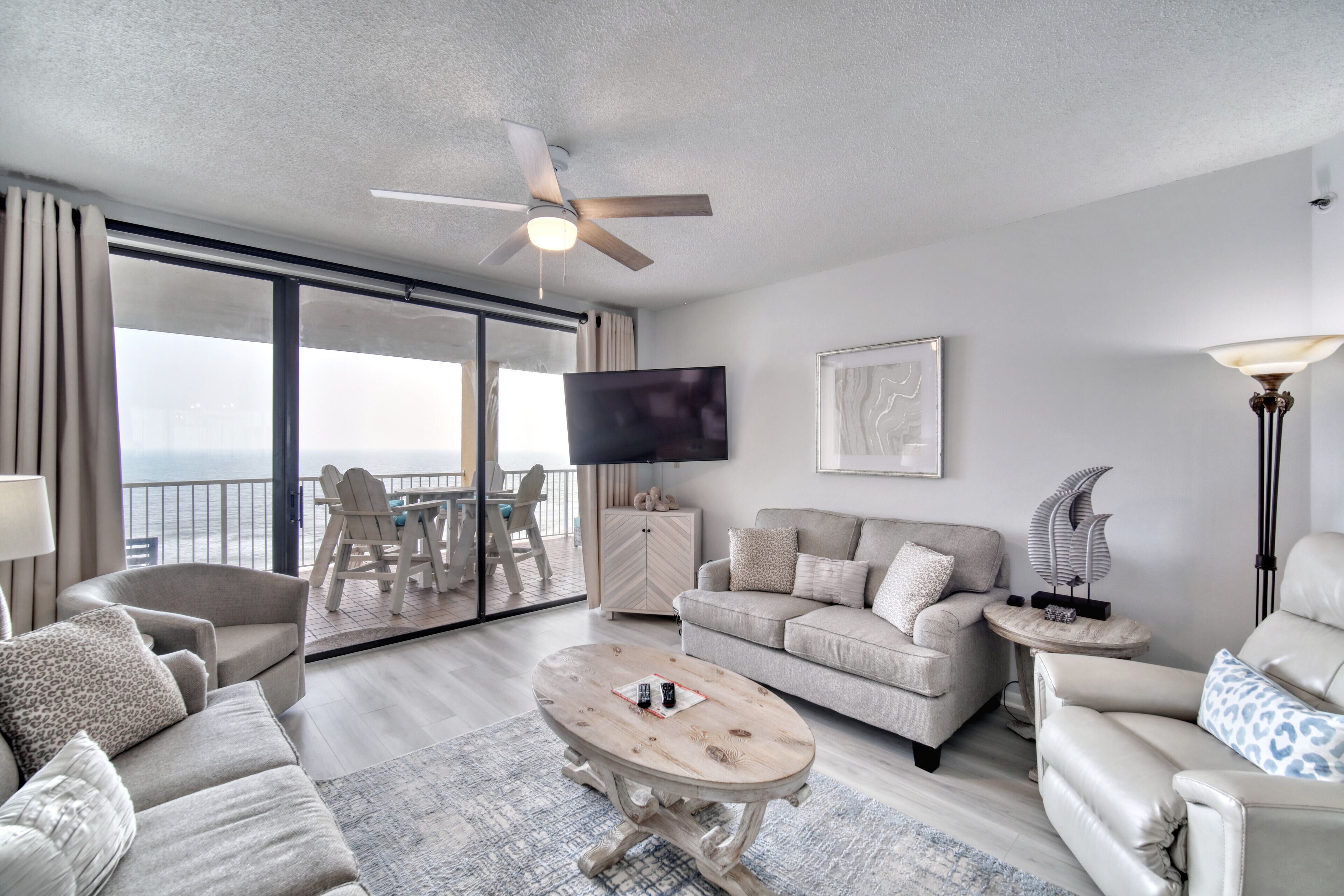 A spacious living area with a ceiling fan, a flat-screen TV, a sofa set, a coffee table, and a view of the outdoors.