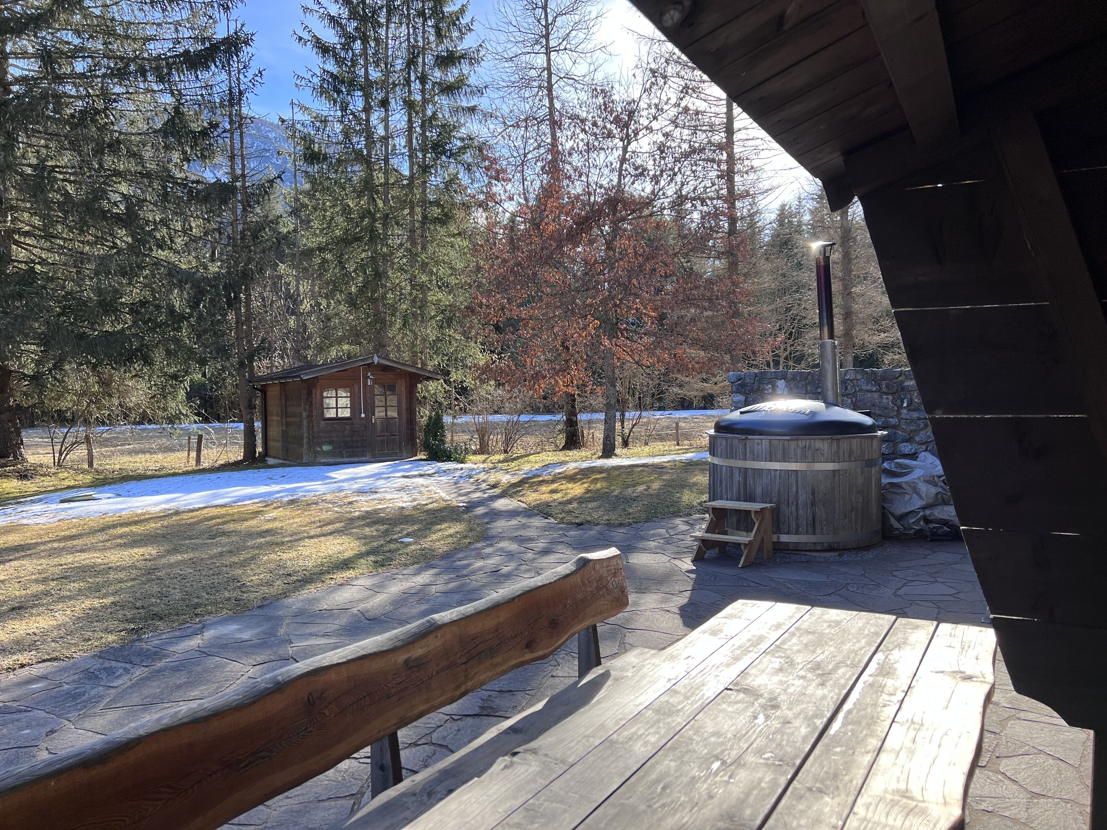 A wooden deck with a bench, a hot tub, and a small cabin in a wooded area with snow on the ground.